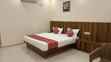 Una habitación de hotel con cabecera de madera, cama con ropa de cama blanca y almohadas con estampado rojo, un pequeño mueble de madera, una silla de mimbre y un juego de mesa en el suelo.