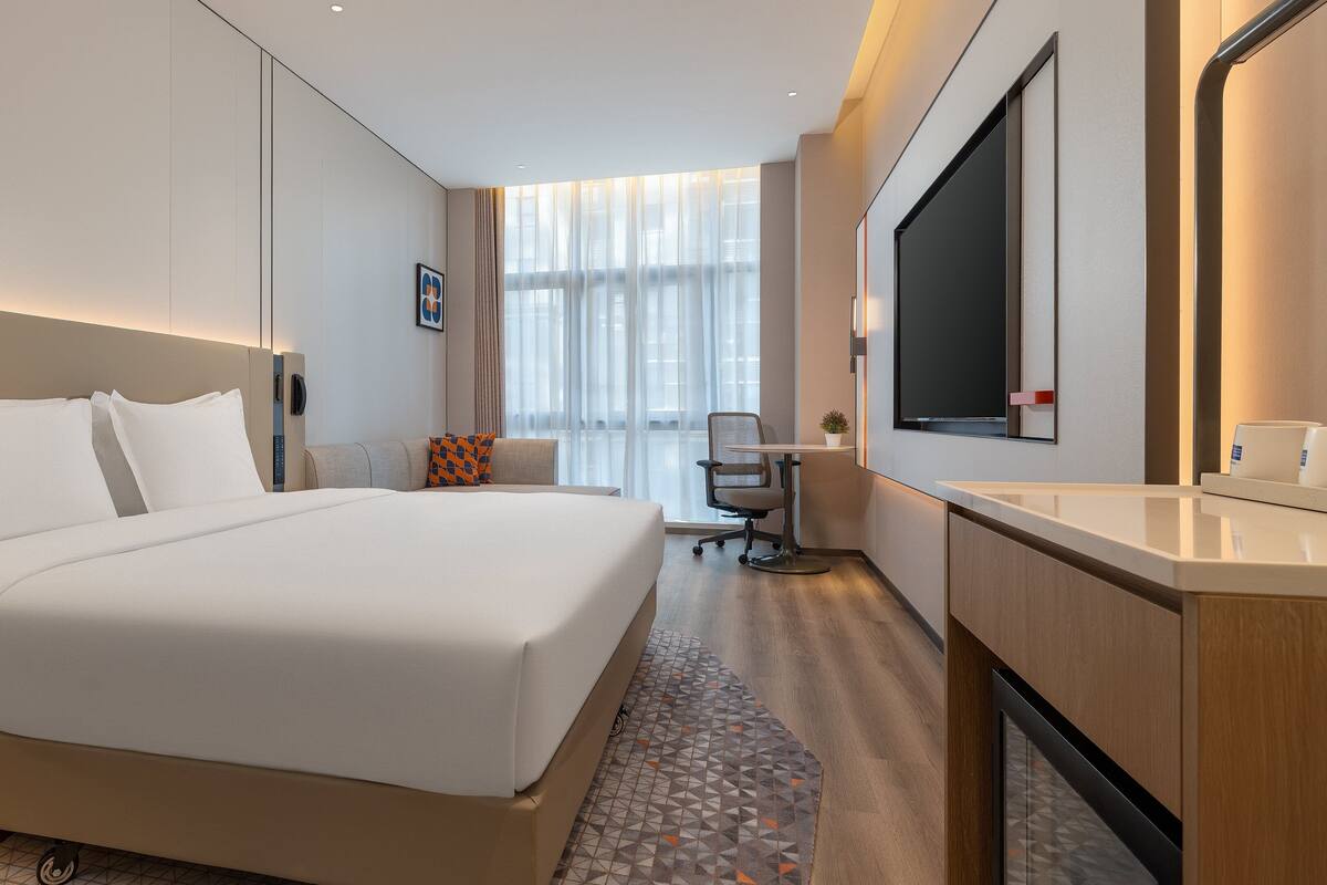 A hotel room with a large bed, a desk with a chair, a TV, and a small plant.