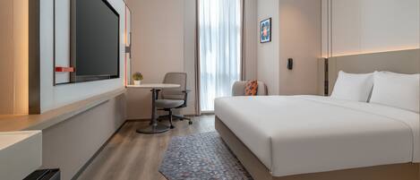 Ein Hotelzimmer mit einem großen Bett, einem Schreibtisch, einem Stuhl, einem kleinen Tisch, einer Topfpflanze und einem Fenster mit Vorhängen.