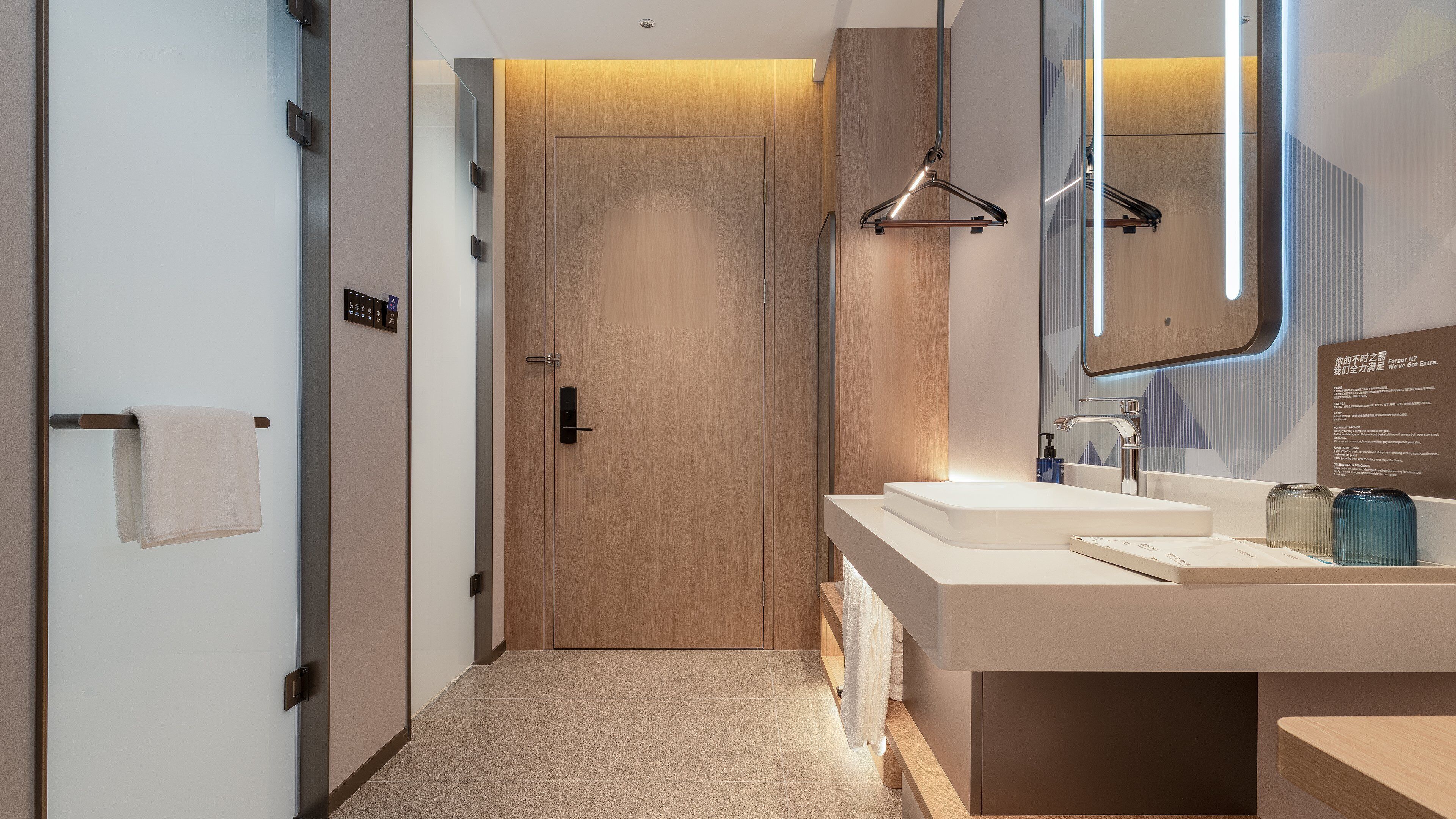 Une salle de bain moderne avec une porte en bois, un lavabo blanc et un miroir doté d’un cadre éclairé.