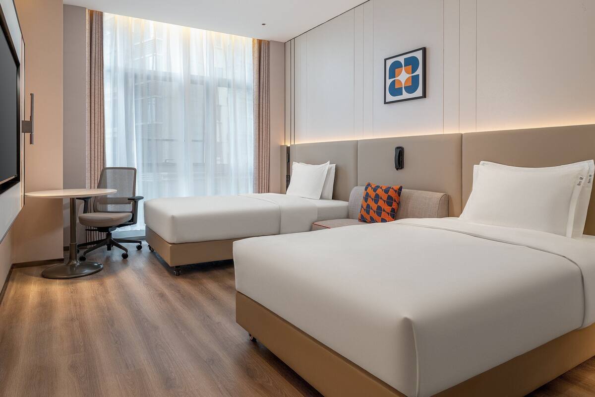 A hotel room with two beds, a desk, a chair, and a large window with sheer curtains.