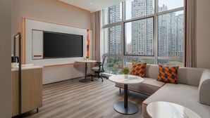 A modern office space with a large flat-screen TV, a round table, a sofa with patterned cushions, and a view of tall buildings through large windows.