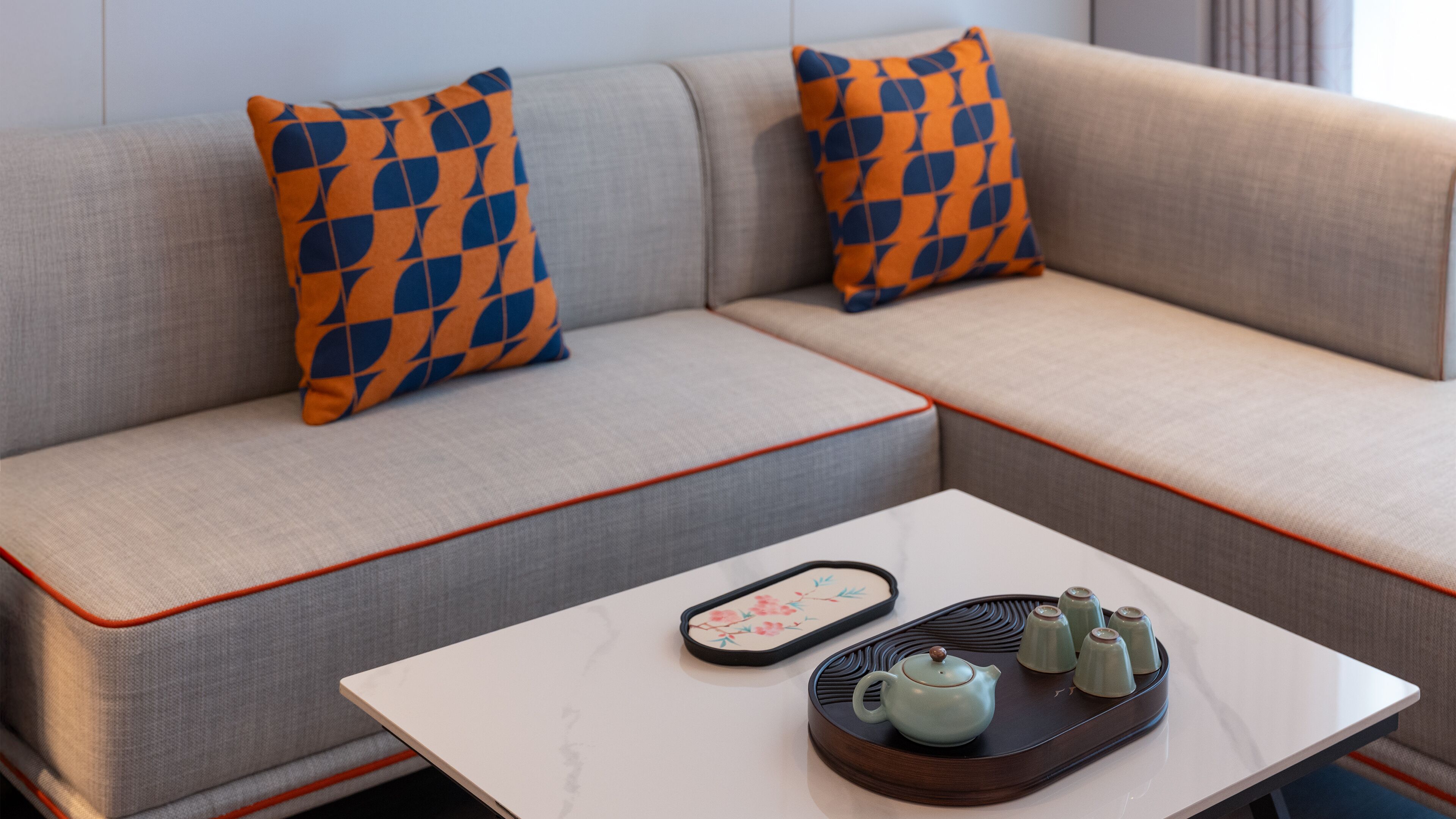 A modern living room with a grey sofa, orange and blue patterned pillows, a white coffee table with a tray of teapots and a remote control.