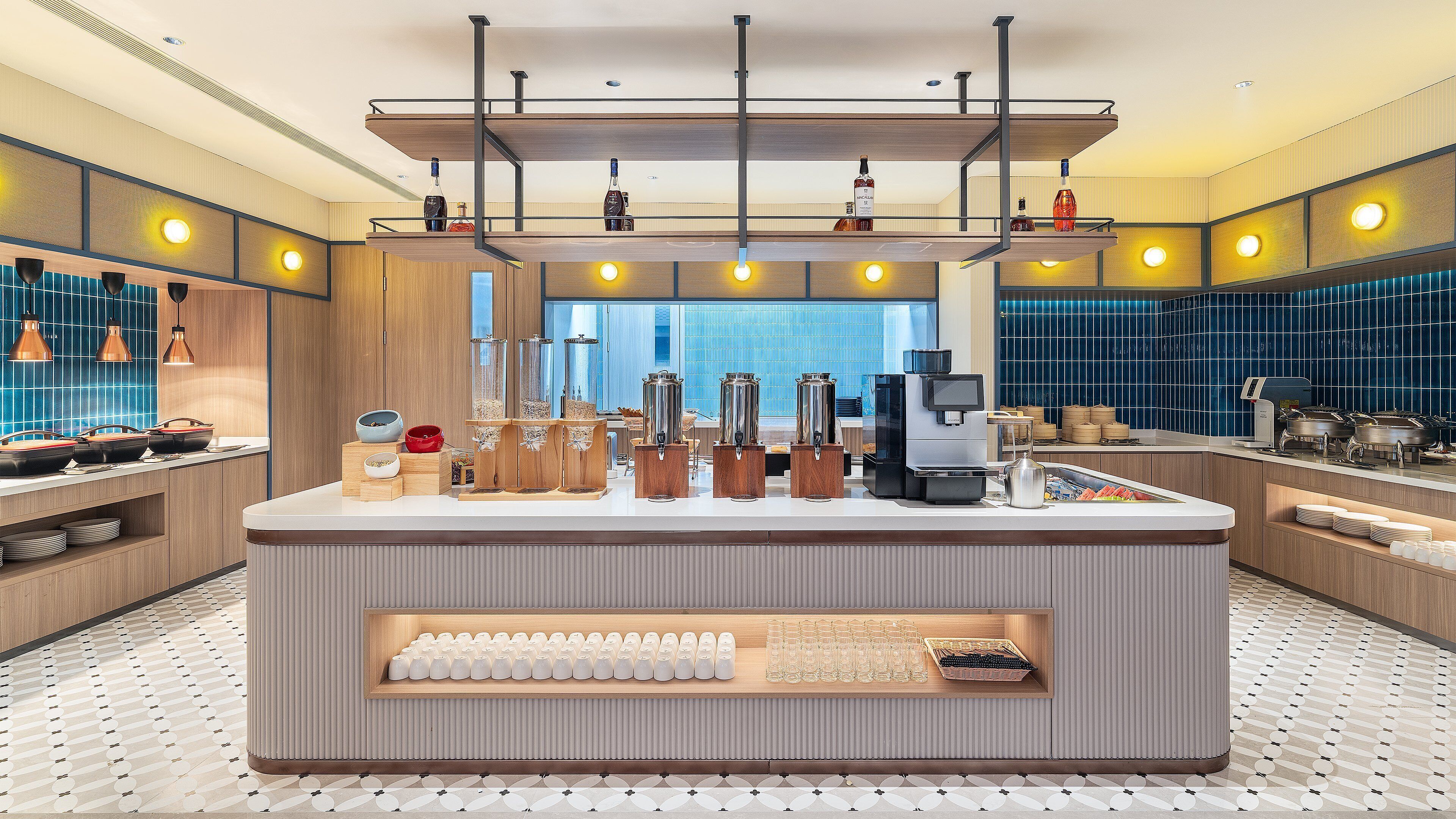 A modern hotel bar with a central counter, multiple beverage dispensers, and a view of the cityscape through large windows.
