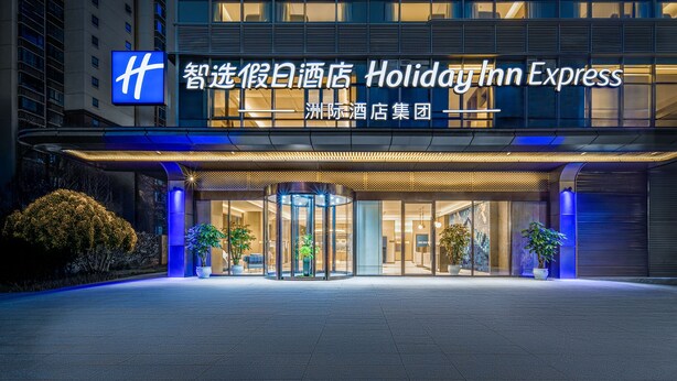 A Holiday Inn Express hotel entrance with illuminated signage, a revolving door, and potted plants. - Holiday Inn Express Kunming Dianchi Rixin Road By Ihg (Kunming)