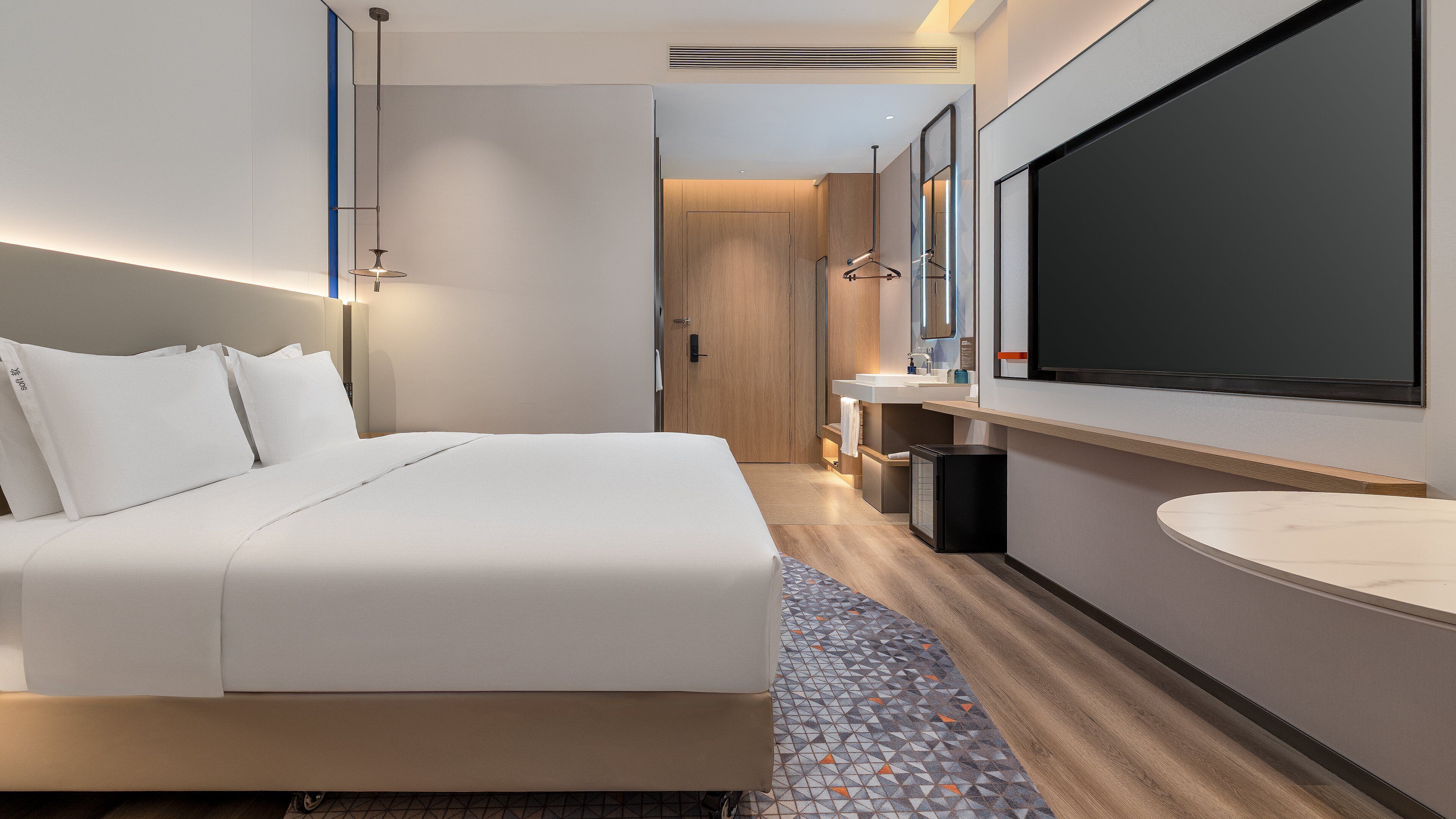 A modern hotel room with a large bed, a bathroom with a mirror and sink, and a flat-screen TV mounted on the wall.