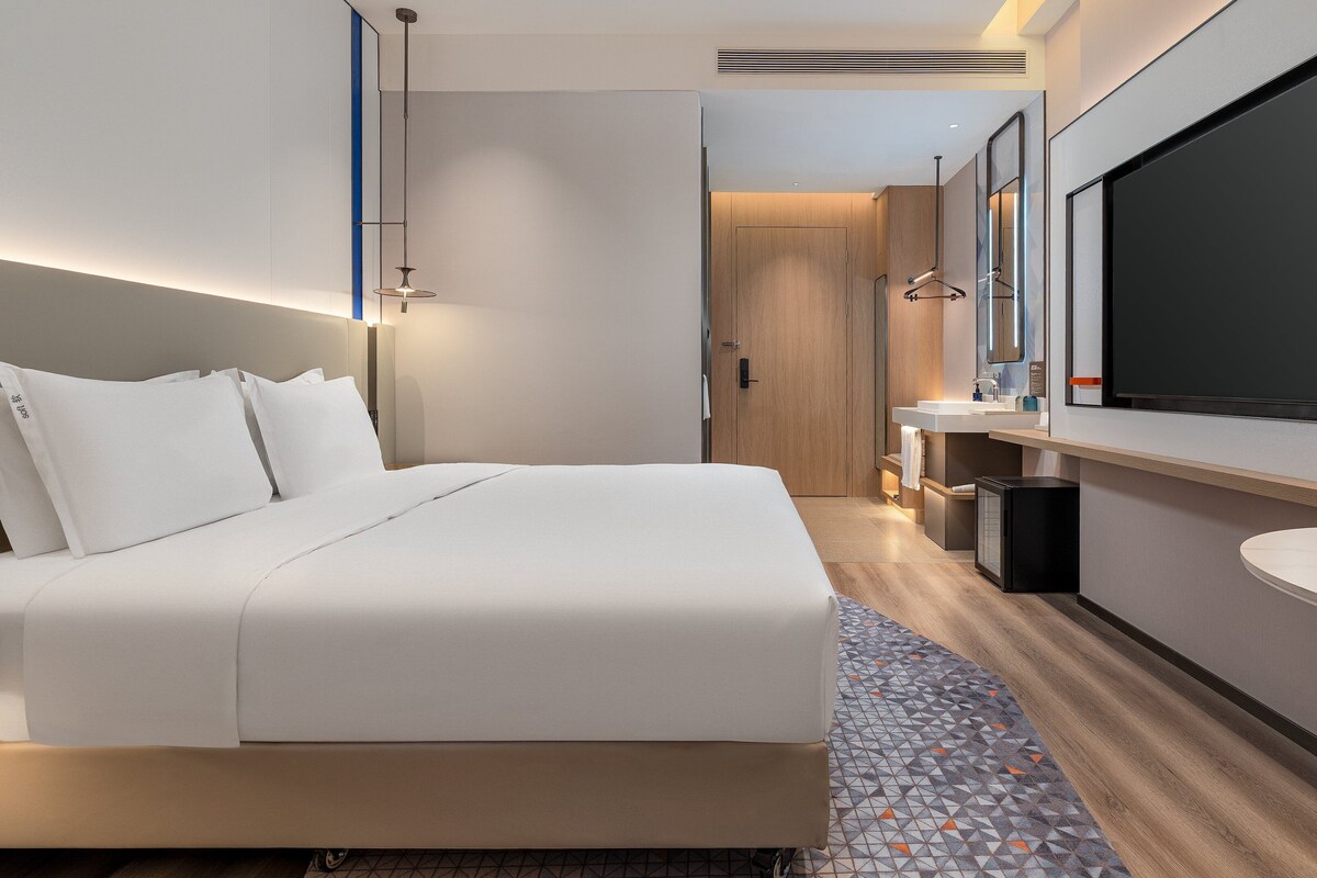 Une chambre d’hôtel moderne dotée d’un grand lit, d’une salle de bain avec un miroir et un lavabo, et d’un téléviseur à écran plat fixé au mur.