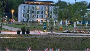 Un bâtiment moderne doté de nombreuses fenêtres, d’un espace extérieur aménagé avec des sièges, ainsi que d’un jardin avec des chaises et des plantes.