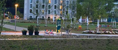 Un edificio moderno con múltiples ventanas, mobiliario exterior y una zona ajardinada con sillas y plantas.