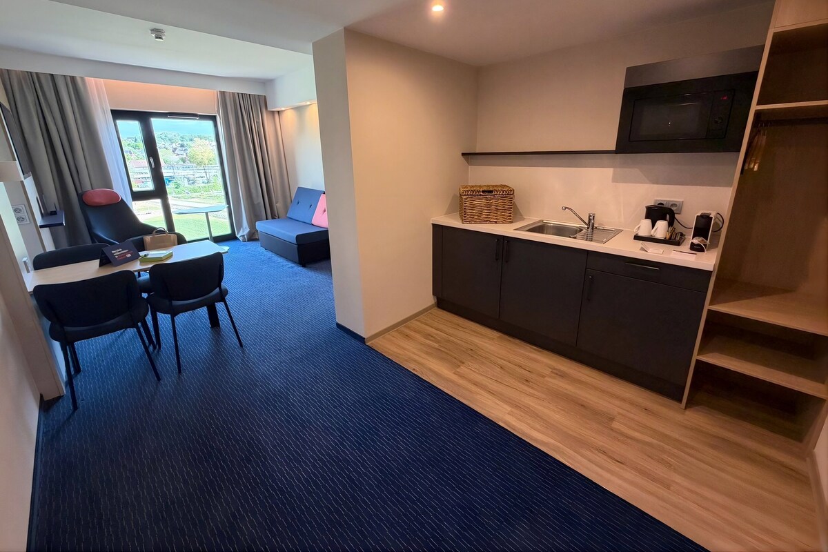 A modern hotel room with a kitchenette, a dining area with a table and chairs, a sofa, and a view through a large window.