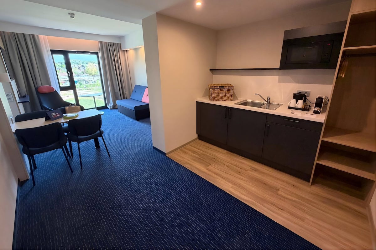 Ett modernt hotellrum med ett litet kök, en matplats med bord och stolar, en soffa och utsikt genom ett stort fönster.