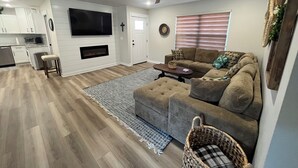 A modern living room with a sectional sofa, a flat-screen TV mounted on the wall, a fireplace, and a dining area with a kitchen in the background.