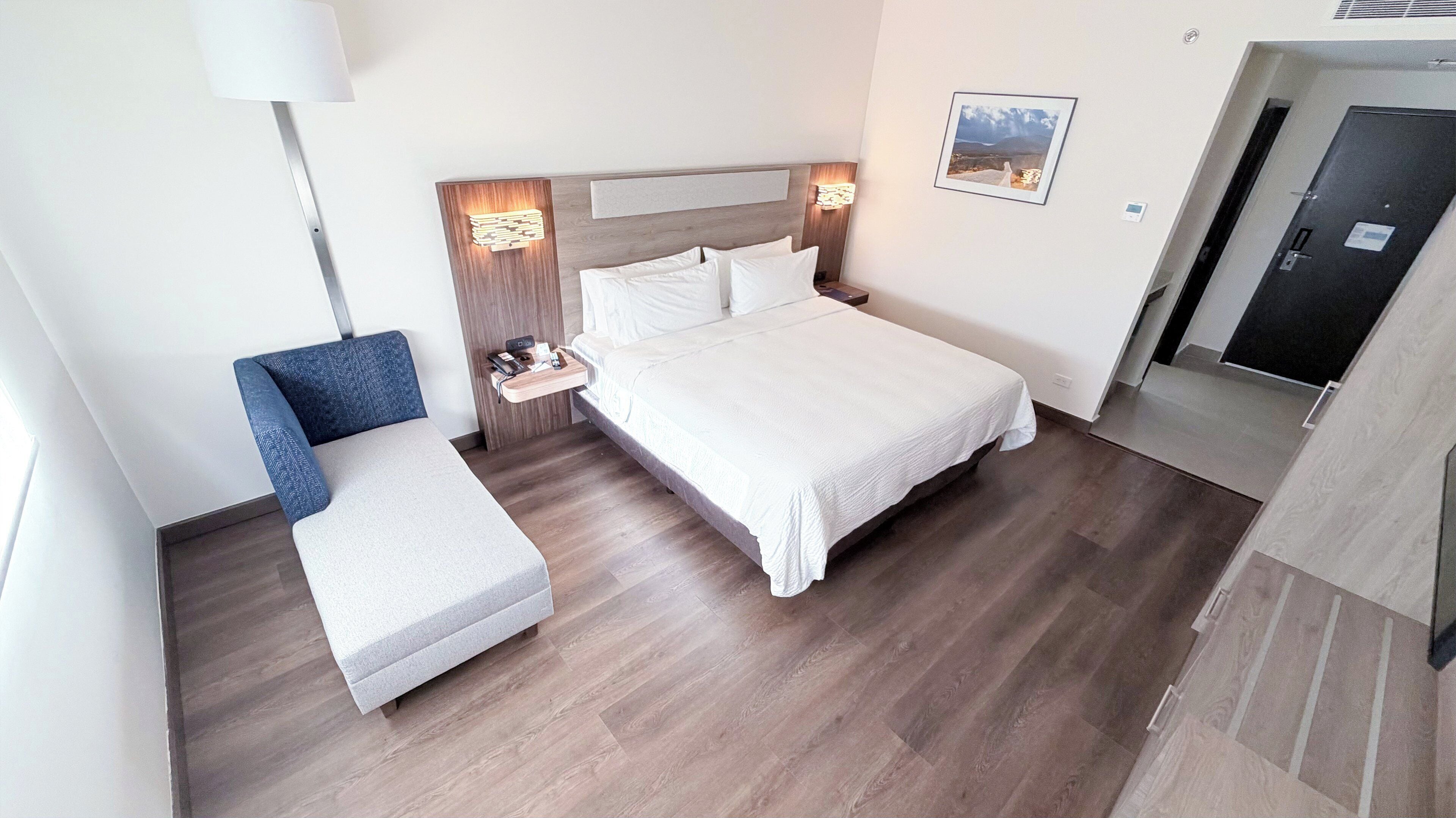 A hotel room with a bed, a bedside table, a chair, a lamp, and a picture on the wall.