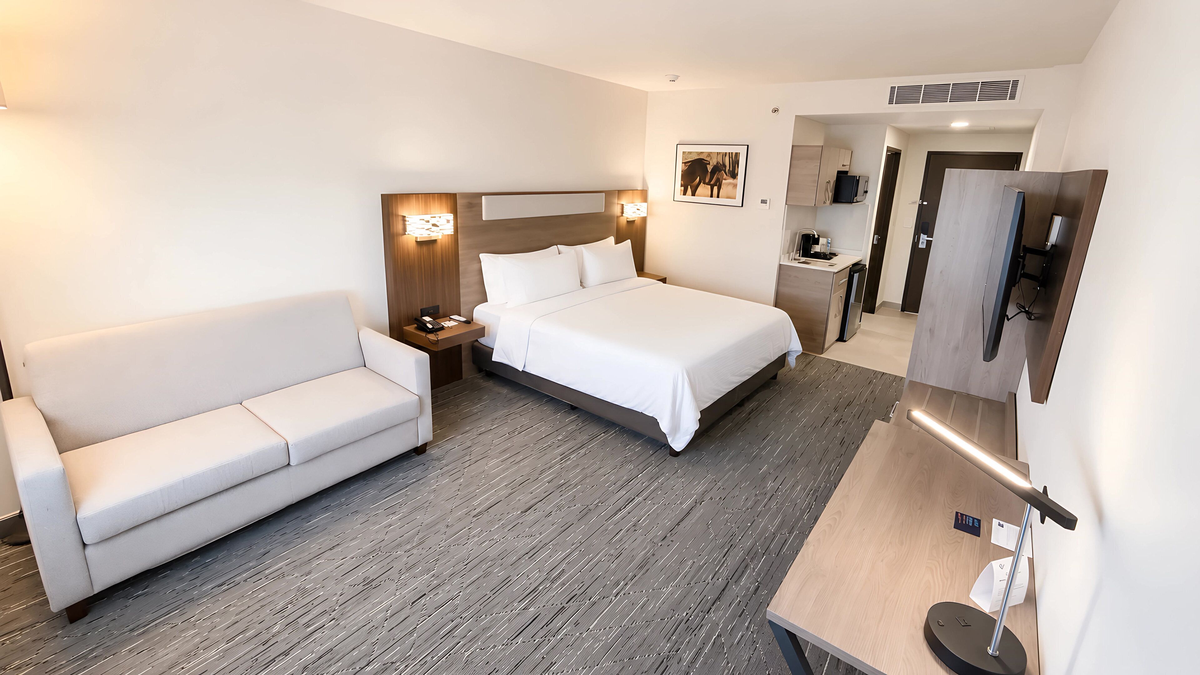 A hotel room with a bed, a sofa, a desk, a mirror, and a small kitchenette.