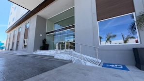 Un bâtiment moderne doté d’une façade en verre, d’une rampe avec un garde-corps et d’un panneau bleu indiquant qu’il est accessible aux personnes à mobilité réduite.