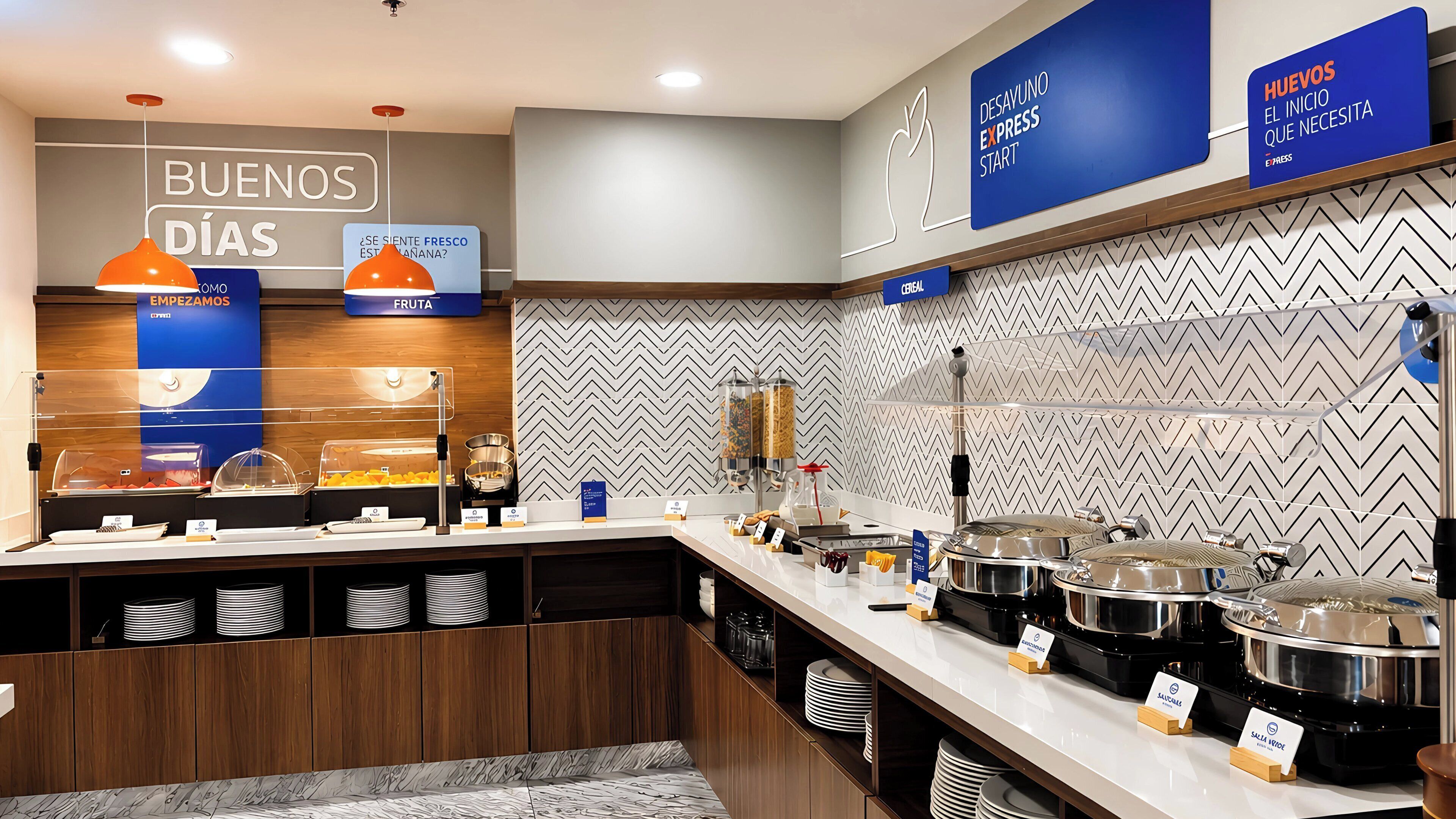 A breakfast buffet area with various food stations, including cereal and fruit options, and a sign indicating the start of the breakfast service.