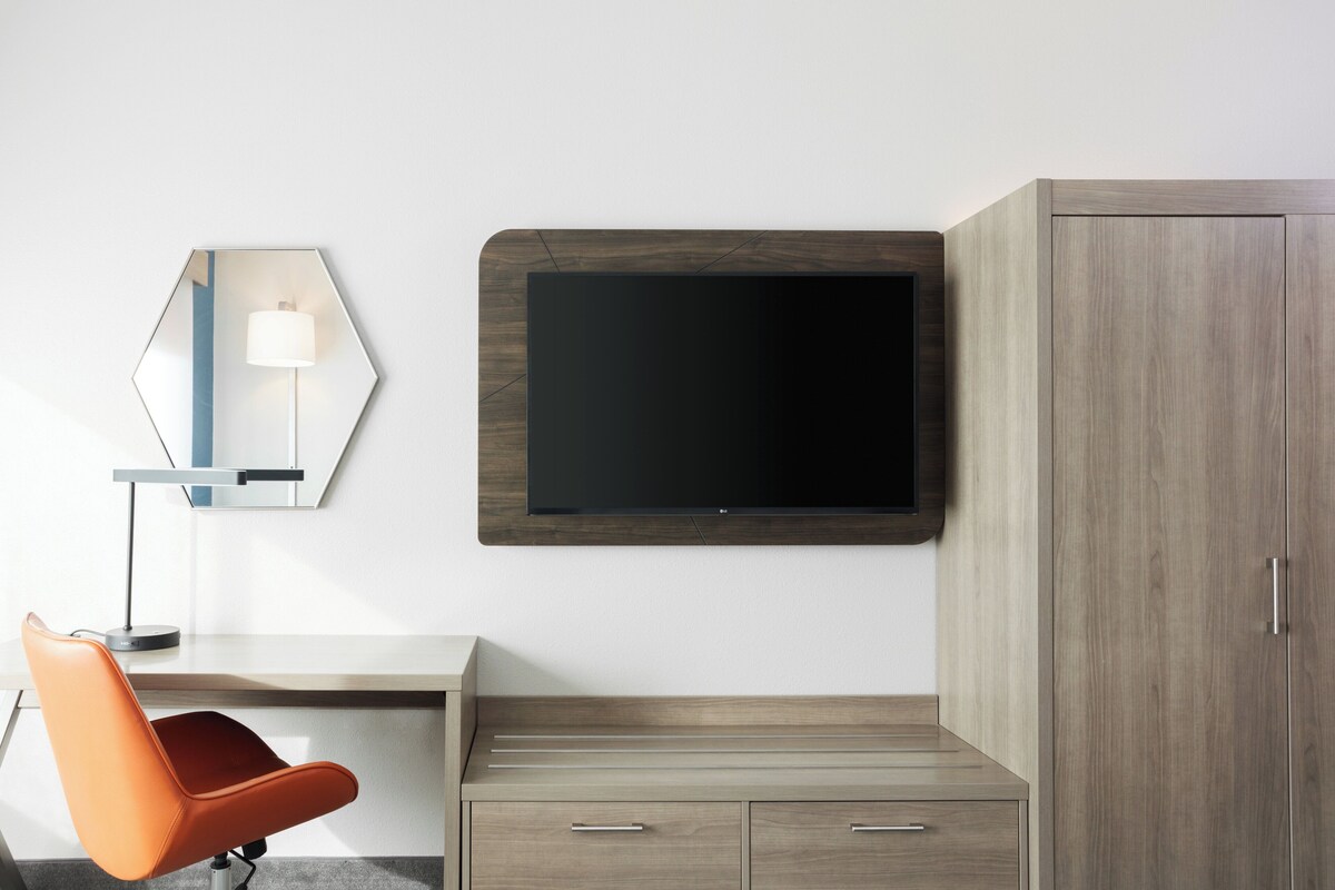 Ein modernes Zimmer mit Flachbildfernseher an der Wand, einem Holzschrank, einem Schreibtisch mit Schubladen und einem orangefarbenen Sessel.