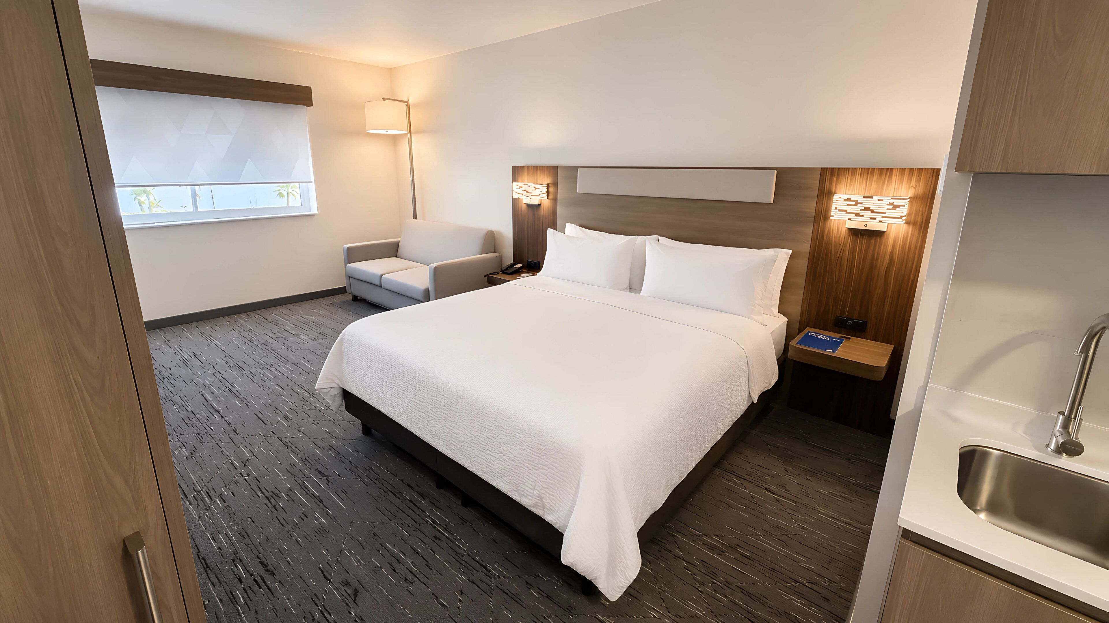 Habitación de hotel con una cama grande, una silla, una mesita de noche con lámpara y vistas al exterior.