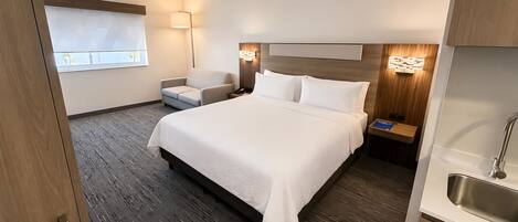 Habitación de hotel con una cama grande, una silla, una mesita de noche con lámpara y vistas al exterior.