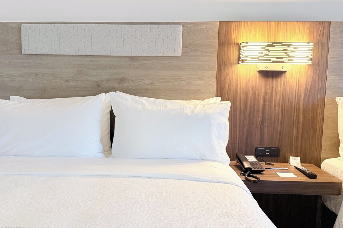 Habitación de hotel con cama, mesita de noche, teléfono, control remoto e iluminación empotrada en la pared.
