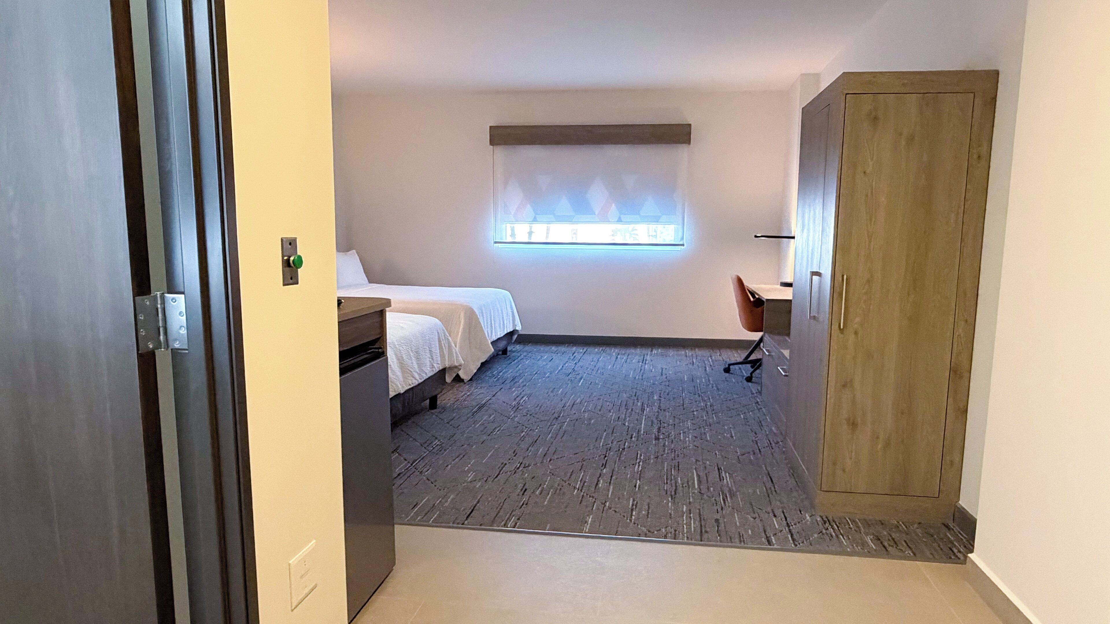 Una habitación de hotel con cama, escritorio, silla, ventana con persianas y puerta.