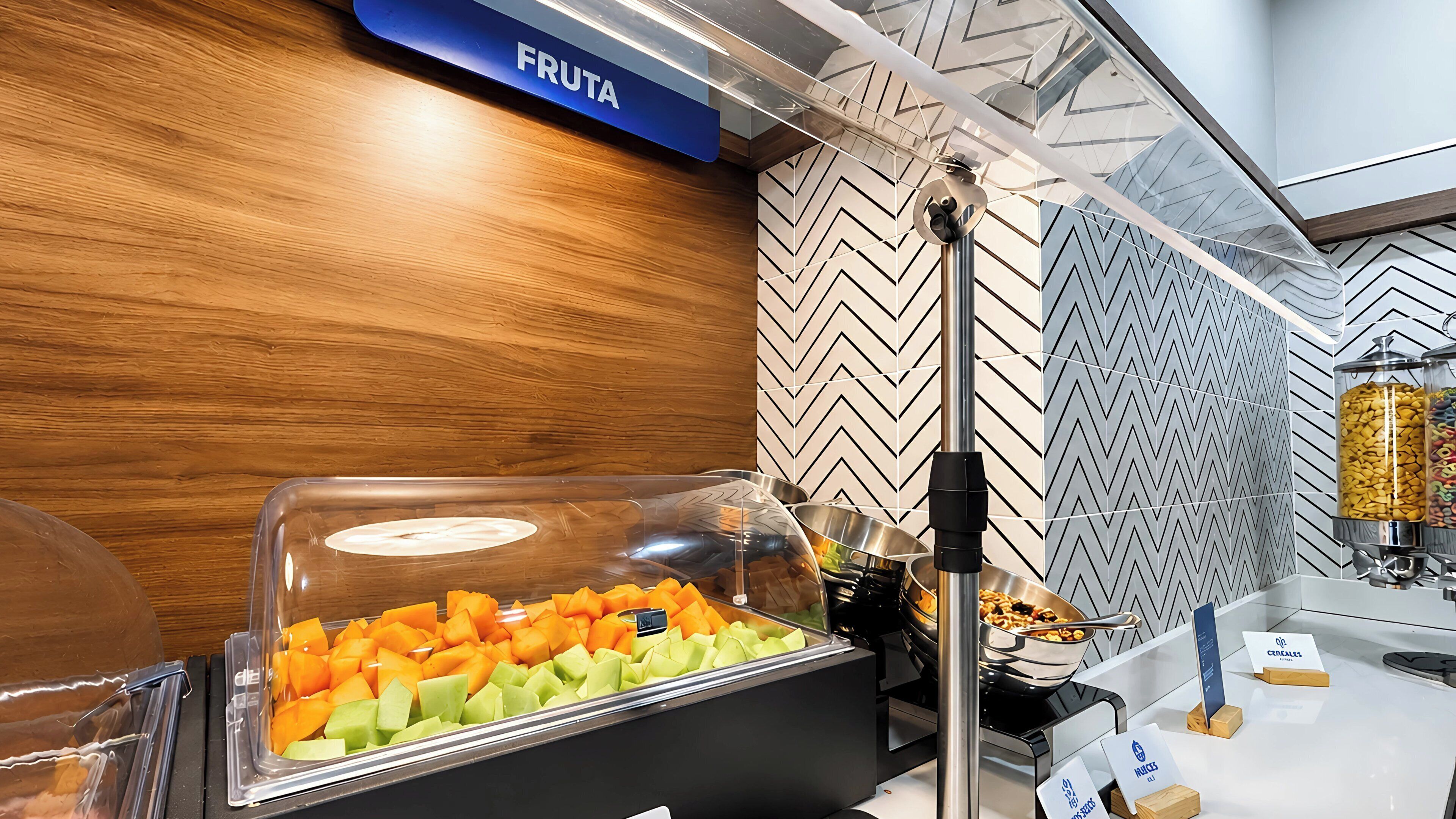 Zona de bufé con un cartel que dice 'FRUTA', una variedad de frutas y una decoración moderna con pared de madera y baldosas estampadas.