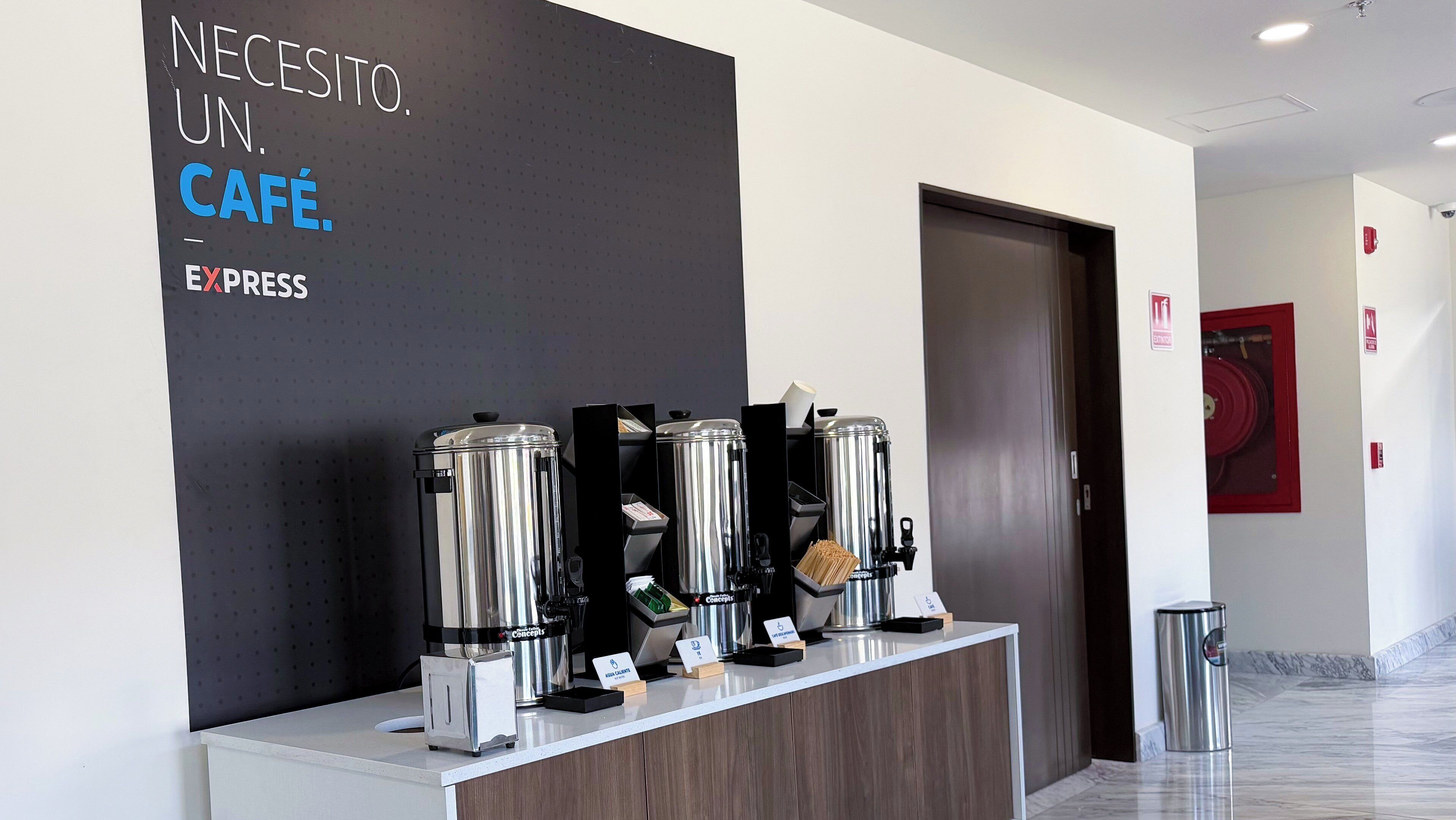 Una estación de café con varias máquinas de espresso, un letrero que dice 'NECESITO UN CAFÉ. EXPRESS' y un interior moderno y limpio.