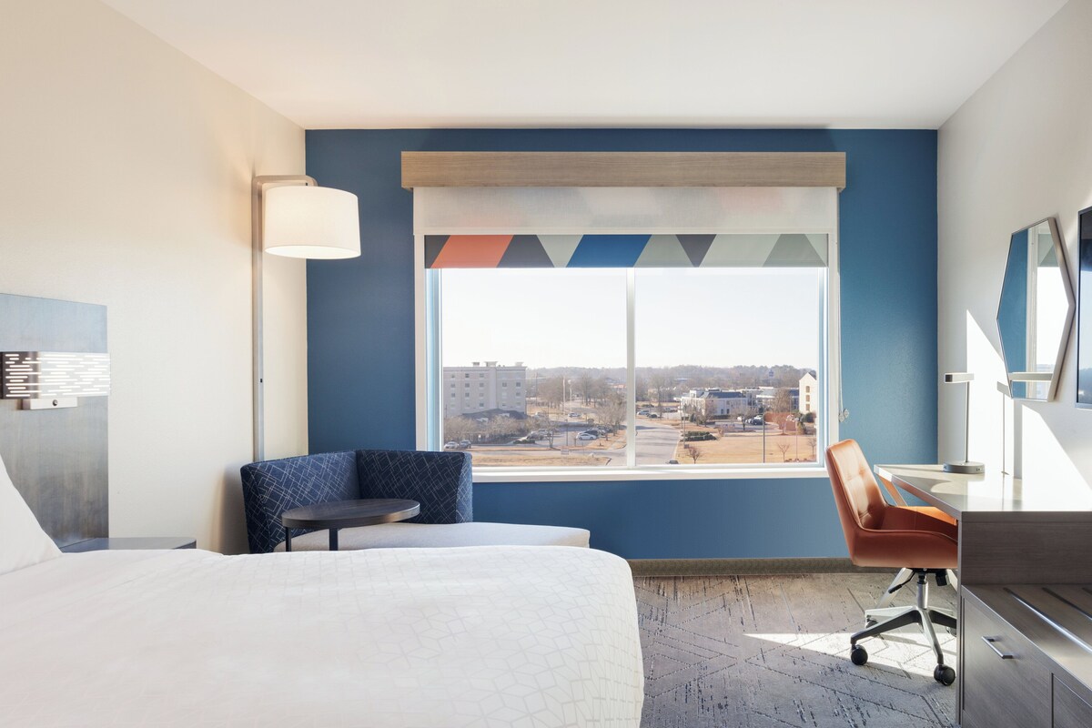 Ein Hotelzimmer mit einem Bett, einem Schreibtisch, einem Stuhl, einer Lampe und einem großen Fenster mit Blick auf Gebäude und einen Parkplatz.