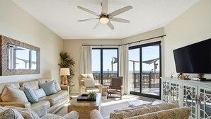 Un salon spacieux comprenant un canapé, des fauteuils, une table basse et un téléviseur. On y trouve également un ventilateur de plafond, une grande fenêtre avec des rideaux et un balcon avec vue.