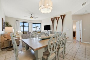 A spacious living area with a wooden dining table, white chairs, a ceiling fan, and a large window with curtains. - Phoenix 10 114 (Orange Beach)