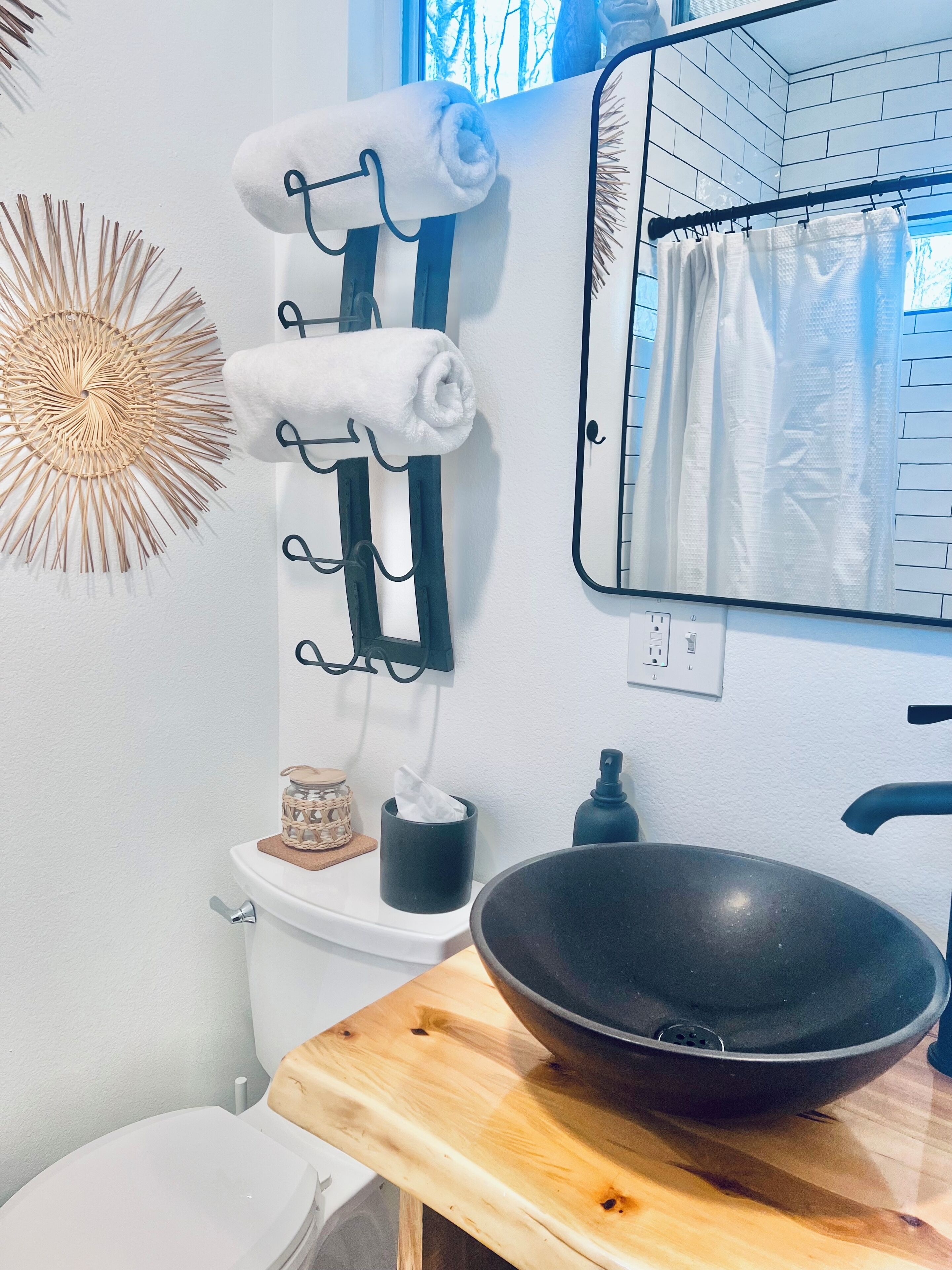 A bathroom with a toilet, a sink with a black basin, a towel rack with towels, a decorative sunburst wall art, and a mirror with a grid pattern.