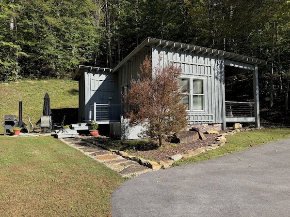 A small cabin with a porch, a grill, and a fire pit in a wooded area.