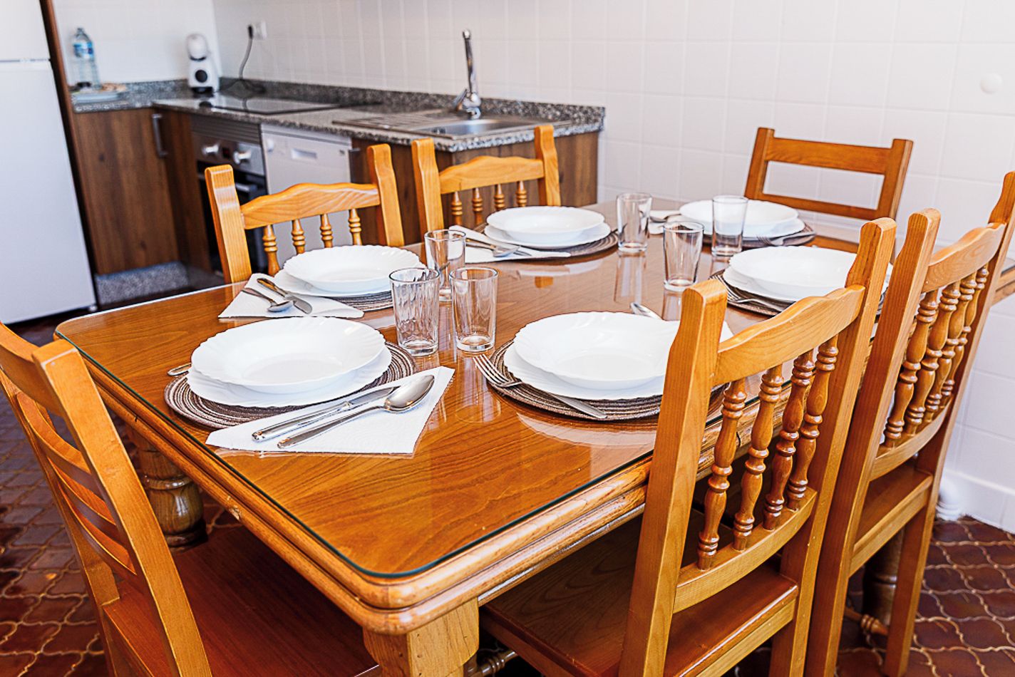Een eettafel gedekt voor zes personen, met houten stoelen, en een keuken in de achtergrond met een betegelde spatwand.