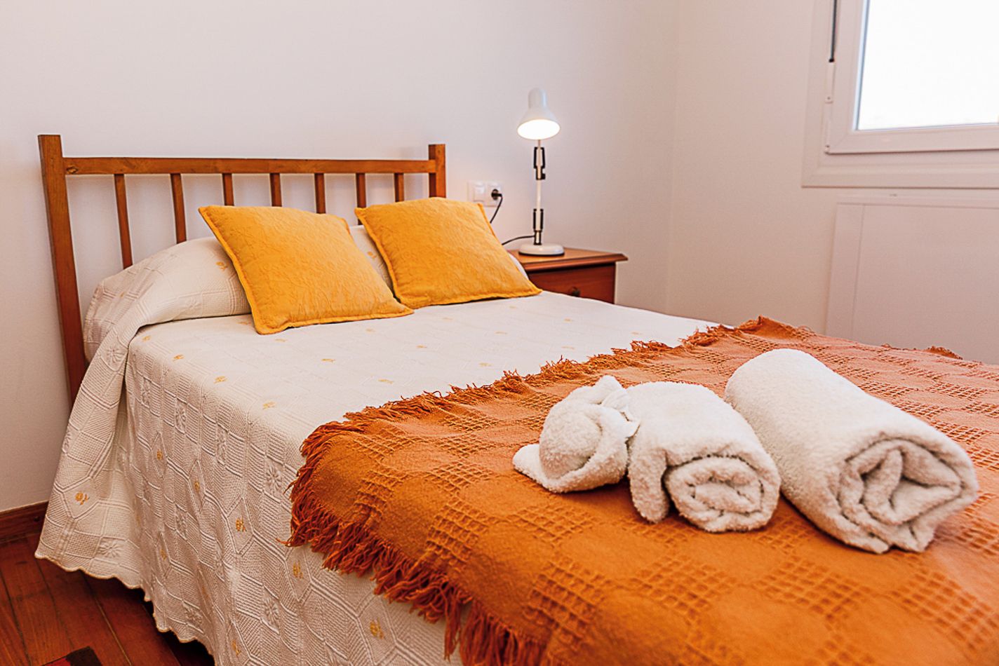 A neatly made bed with a white and orange bedspread, two pillows, and a rolled towel on the side.