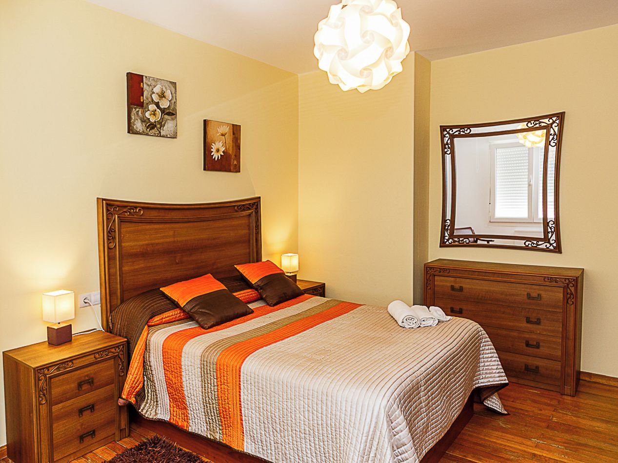 Een netjes opgemaakt bed met een gestreept dekbed, twee nachtkastjes met lampen, een dressoir en een spiegel aan de muur.