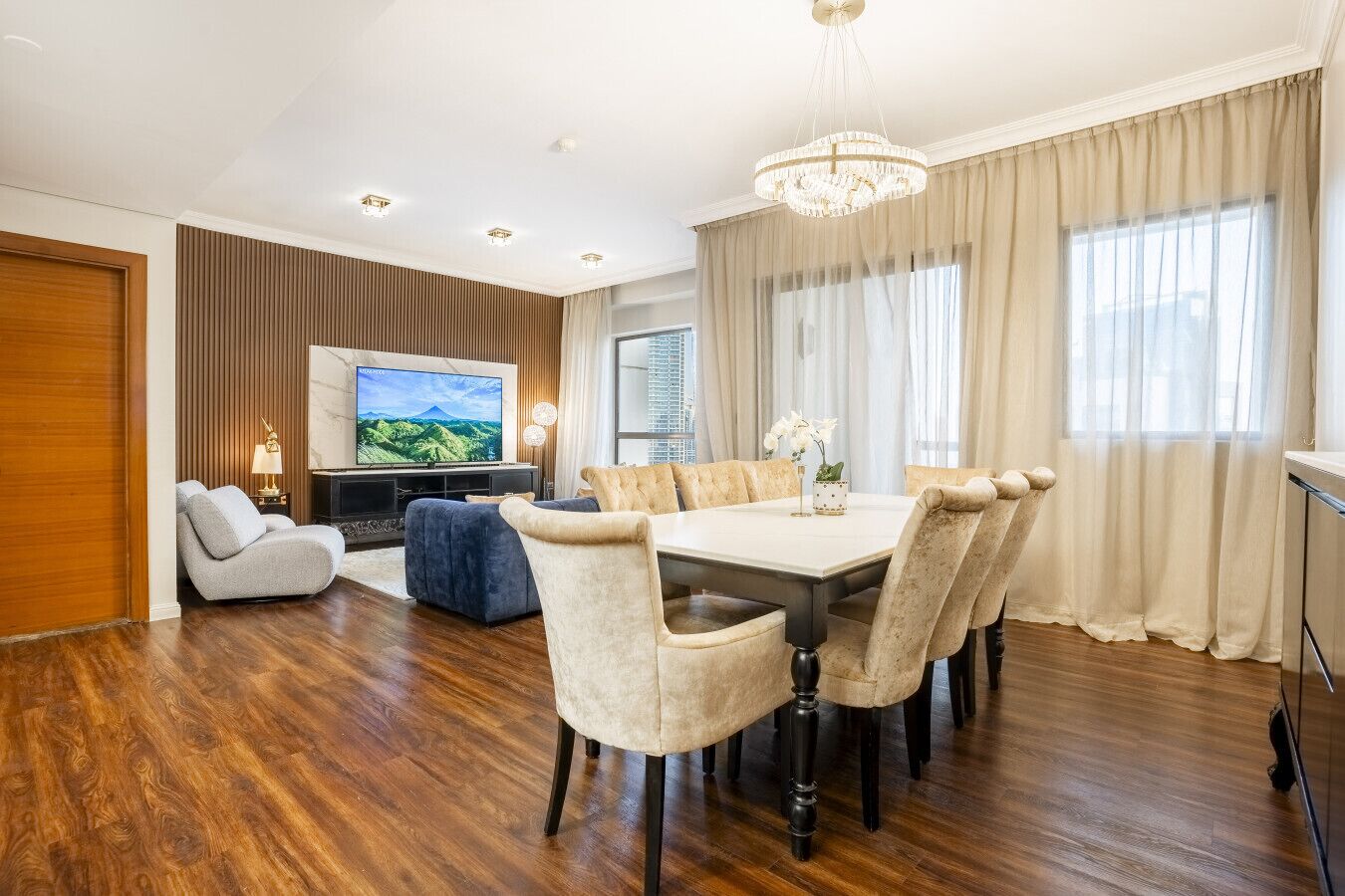 Ein modernes Wohnzimmer mit Holzboden, einem großen Fernseher, einem Esstisch mit Stühlen und einer Chandelieber.