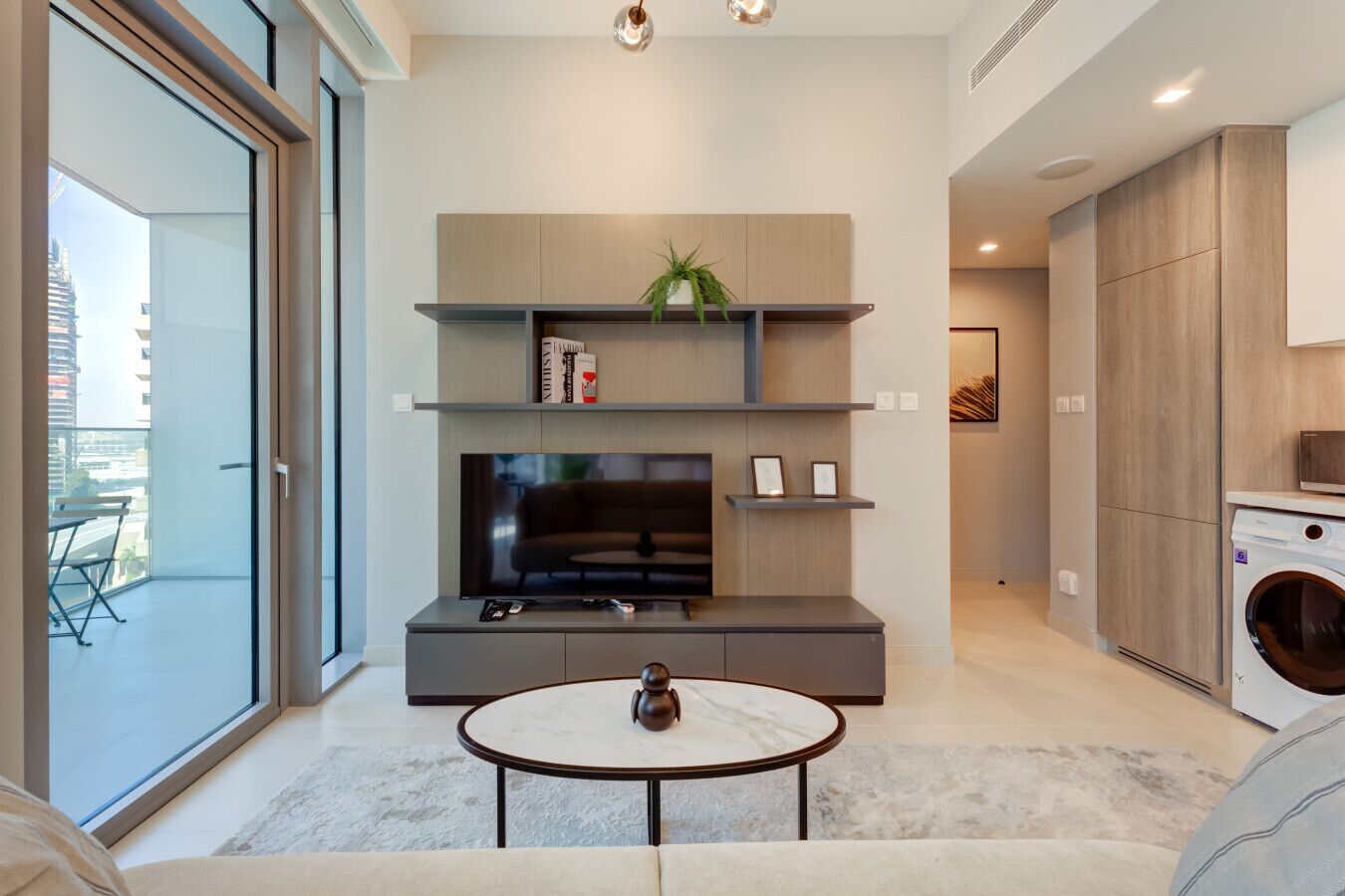 A modern living room with a flat-screen TV, a coffee table, and a sofa. There is a balcony with a view of the city.