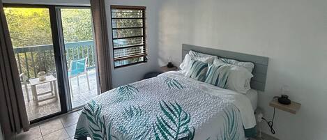 Un dormitorio con cama, ventilador de techo, ventana con persianas, balcón y una pequeña mesa.