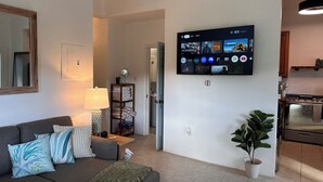 Un soggiorno con televisione a schermo piatto montata a parete che mostra diverse piattaforme di streaming, un divano grigio con cuscini decorativi, un tavolino con una lampada e una pianta in vaso.