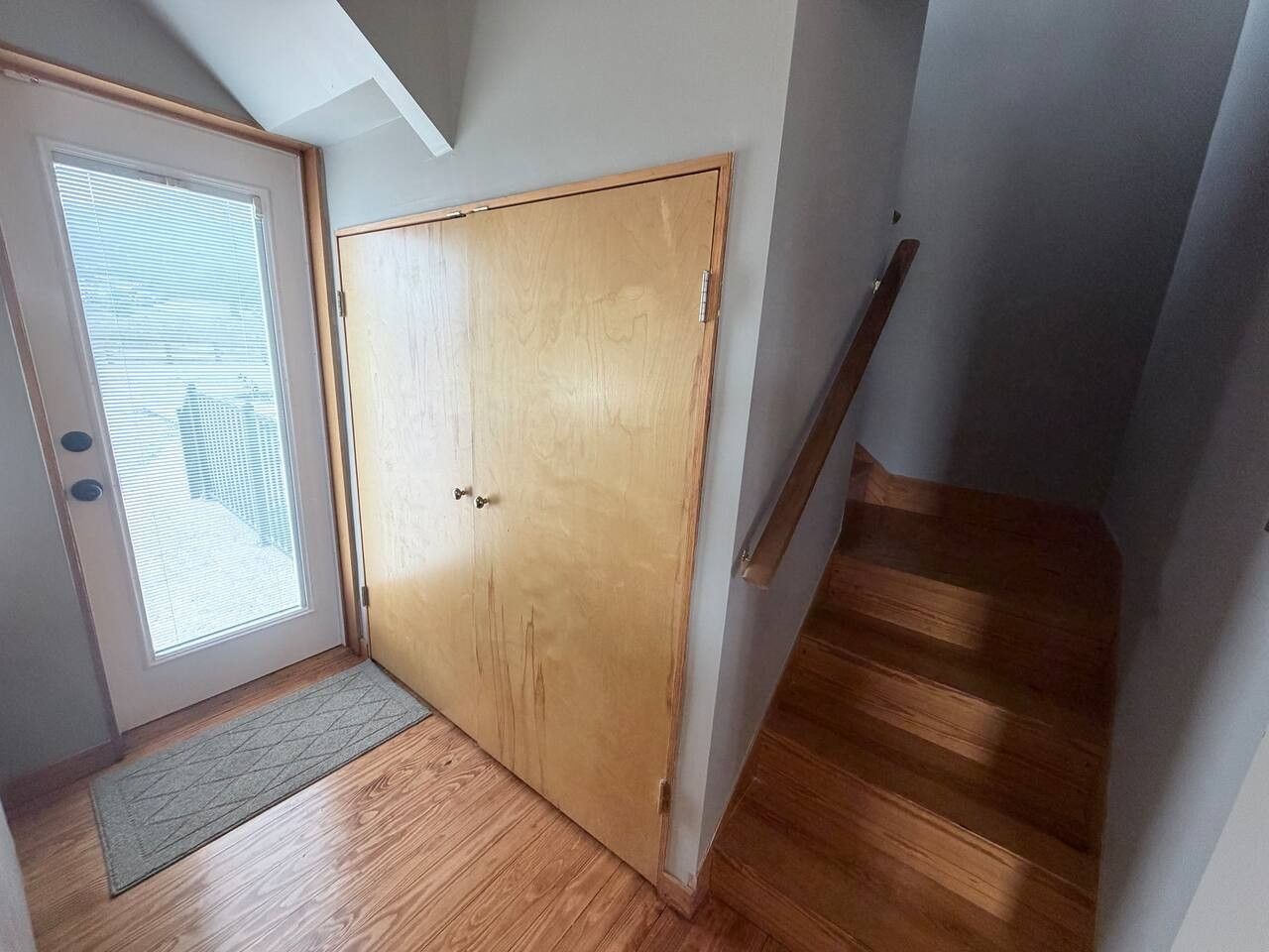 A room with a wooden door, a window with blinds, and a wooden staircase.