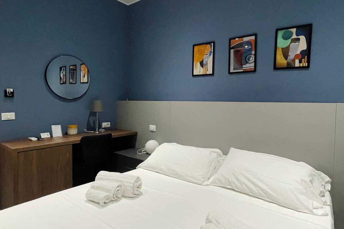 A hotel room with a bed, a desk, a chair, a lamp, and three paintings on the wall.
