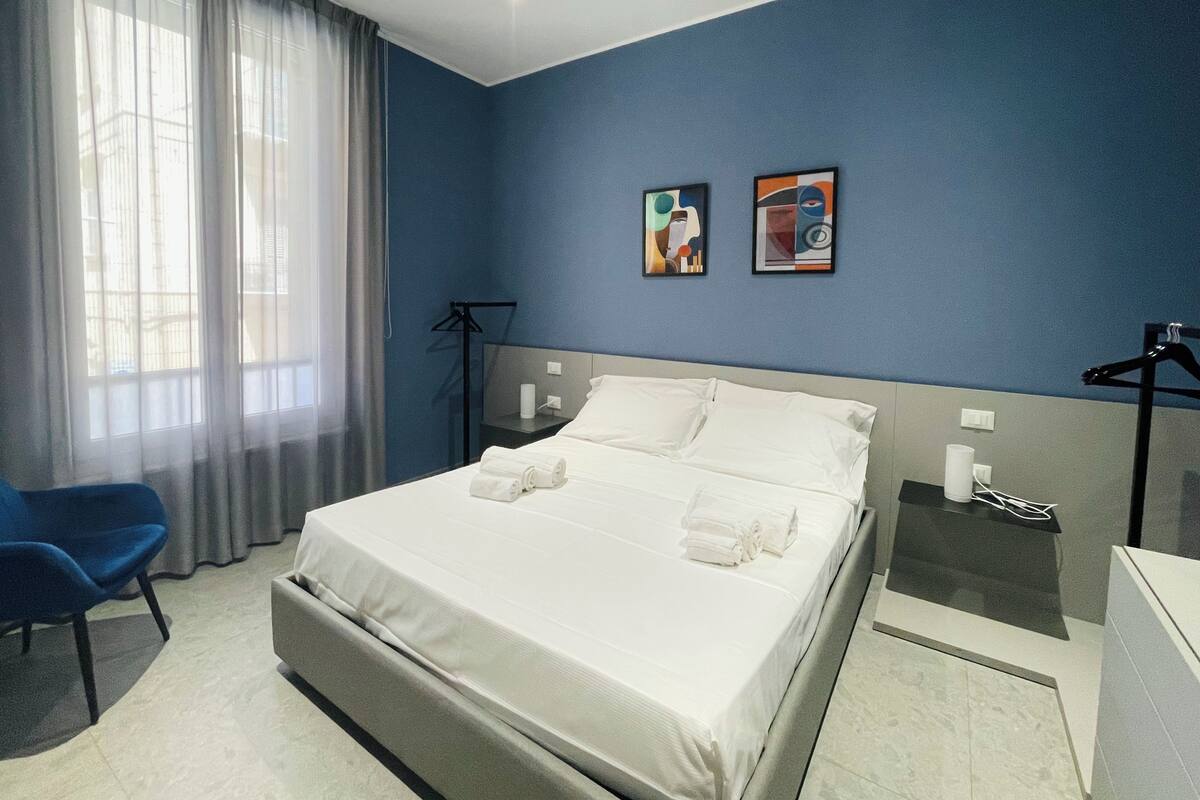 A hotel room with a bed, a chair, a desk, and two paintings on the wall.
