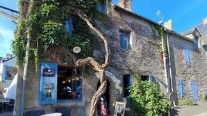 A stone building with ivy-covered walls and a tree growing through the entrance. There are outdoor tables and chairs in front.