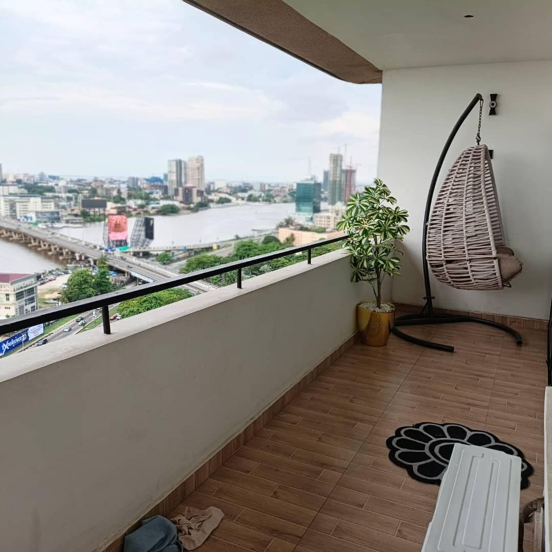 A balcony with a view of a cityscape, a hanging chair, a potted plant, and a patterned mat.