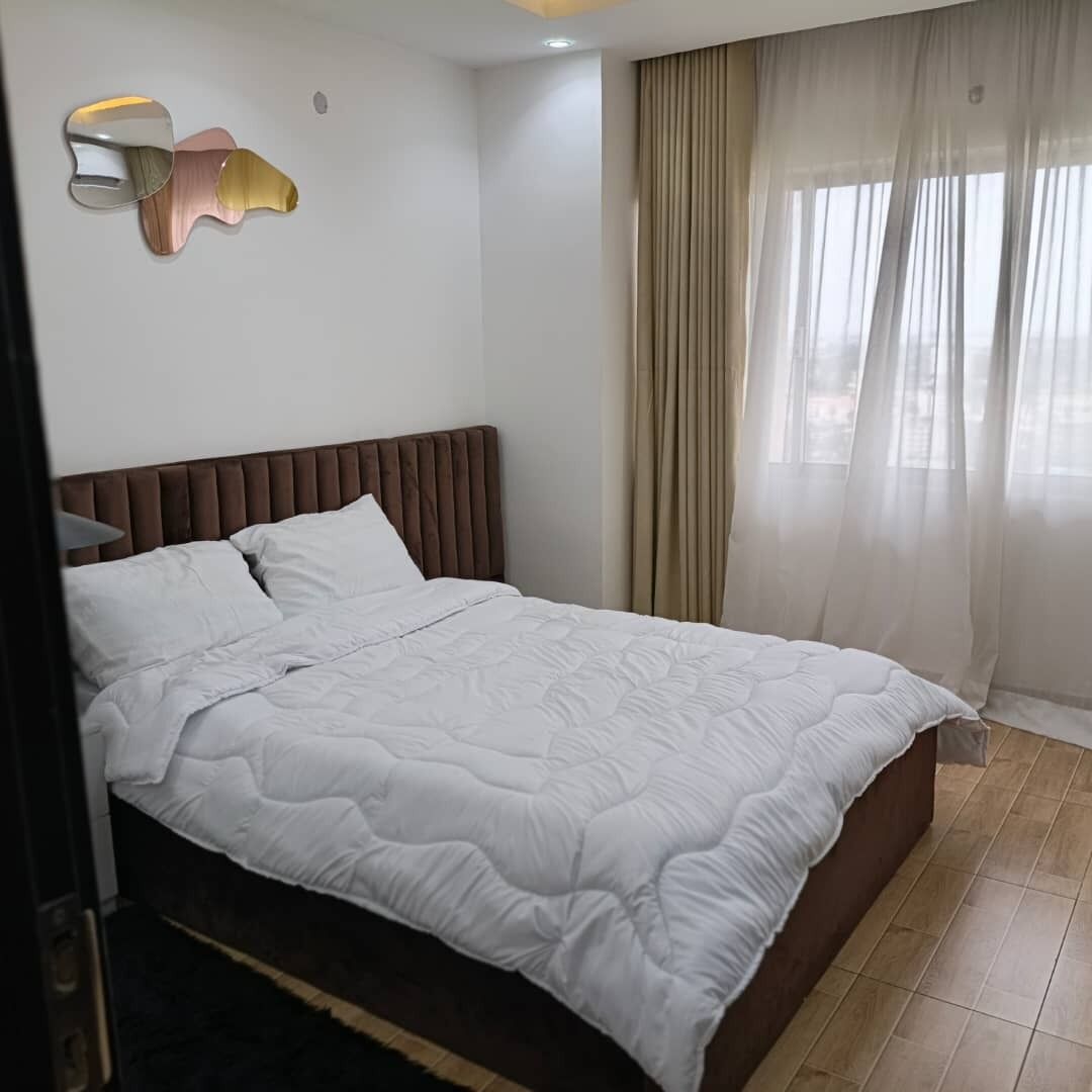 Una cama bien tendida con un cabecero marrón, una ventana con cortinas traslúcidas y una lámpara fijada a la pared.