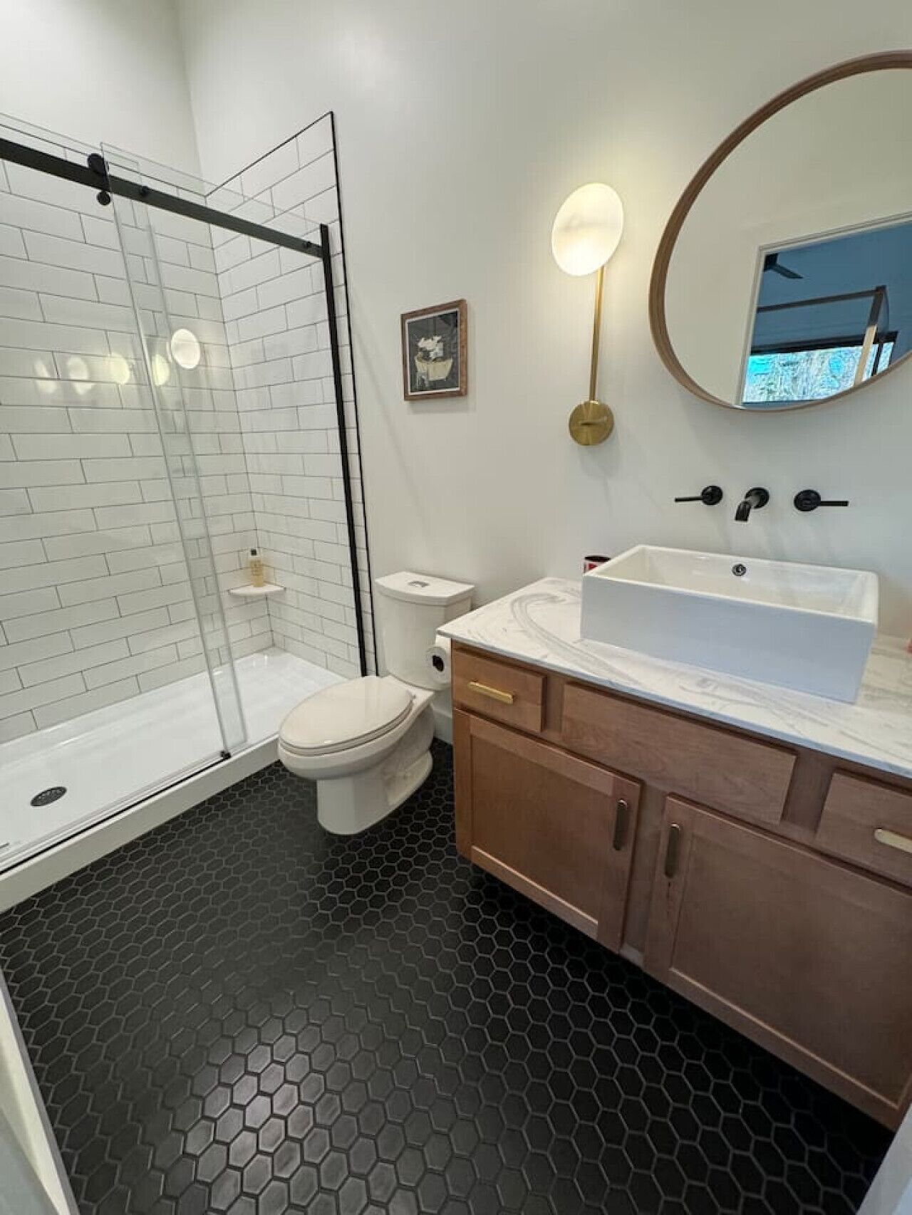 A modern bathroom with a glass shower enclosure, a white toilet, a large sink with a marble countertop, wooden cabinets, and a hexagonal floor pattern.