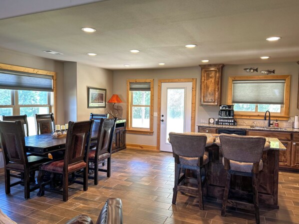 A spacious kitchen with wooden cabinets, a dining area with dark wooden chairs, and a view of the outdoors through large windows. - Ozark Getaway at The White River! (Flippin)