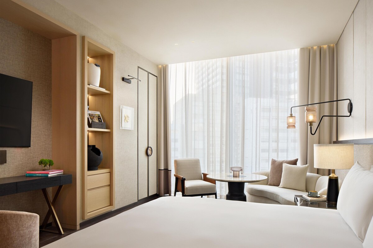 Habitación de hotel con cama, escritorio, silla, mesita, lámpara, televisor y ventana con cortinas.