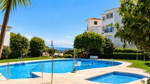 Una piscina con dos zonas separadas, rodeada de palmeras y un edificio con balcones.