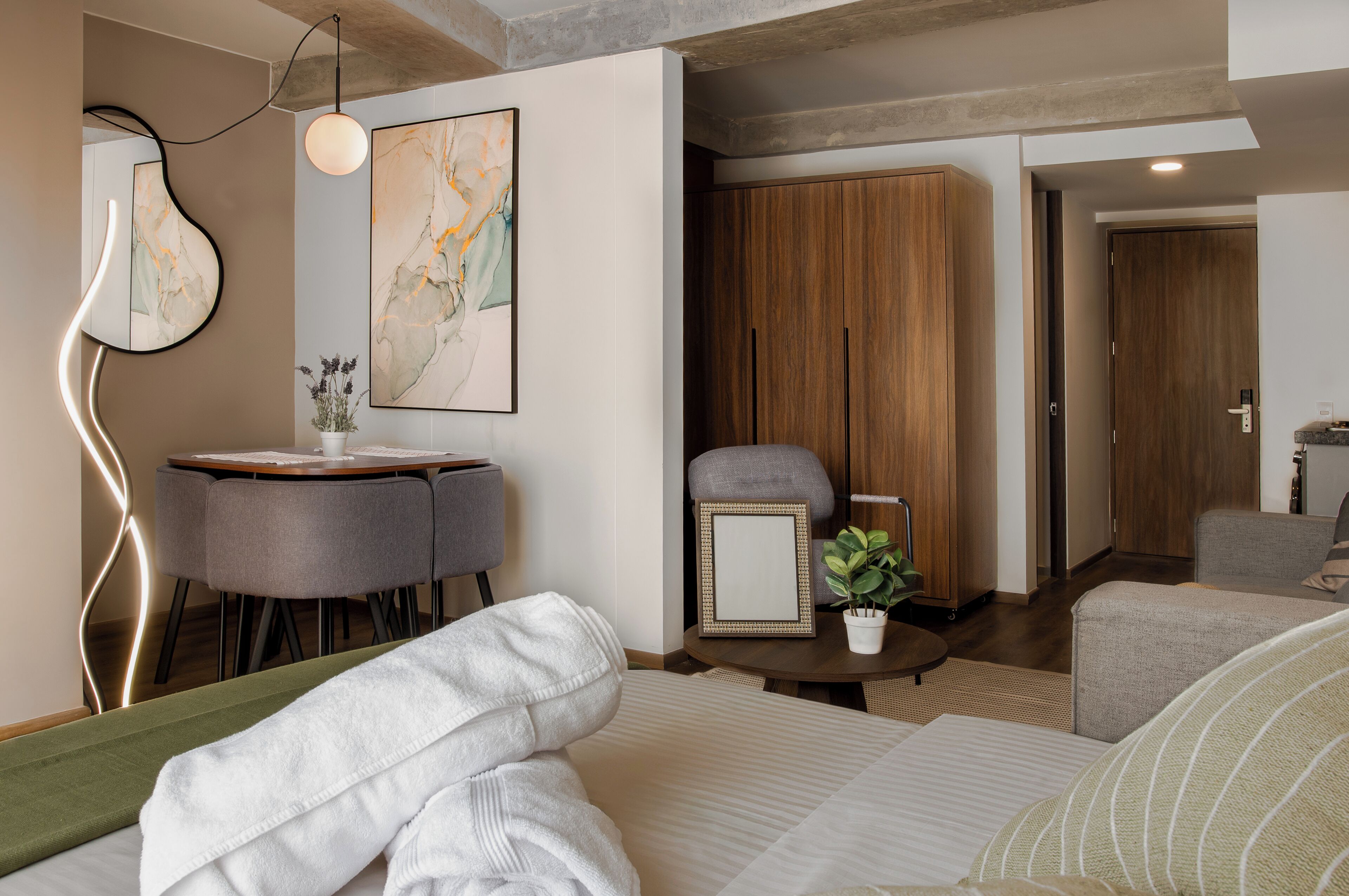 Una habitación de hotel moderna con cama, una pequeña mesa redonda con una planta, un sillón gris y un armario de madera.